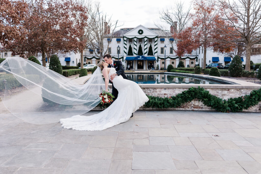 Groom dips bride with veil flying for portrait in front of five star Williamsburg wedding venue