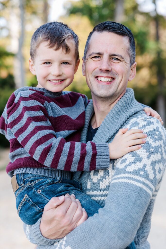 father and son smile at camera while father holds son on beach during session by photographer Williamsburg, VA
