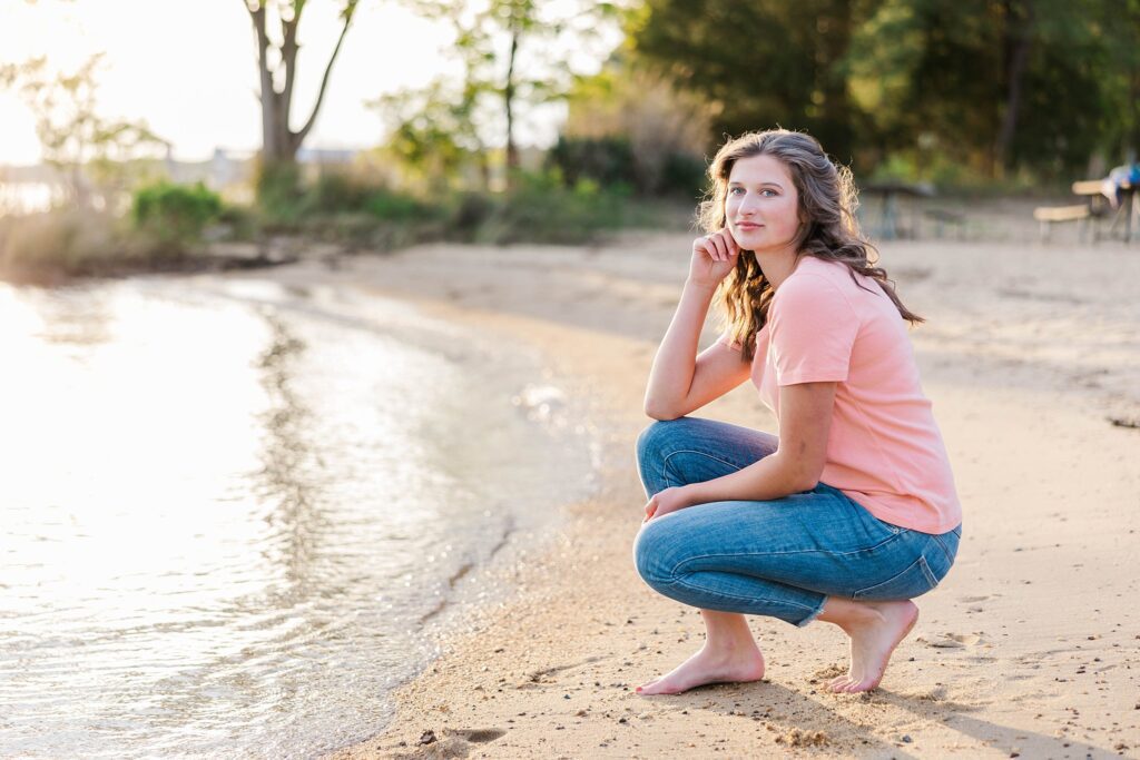 senior in pink shirt and jeans squats by water on James River for senior session in Williamsburg, VA
