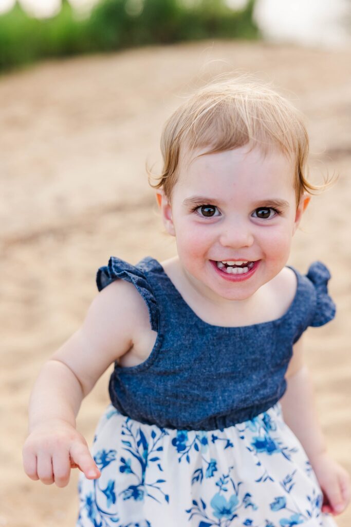 toddler smiles in the sand wearing blue dress
