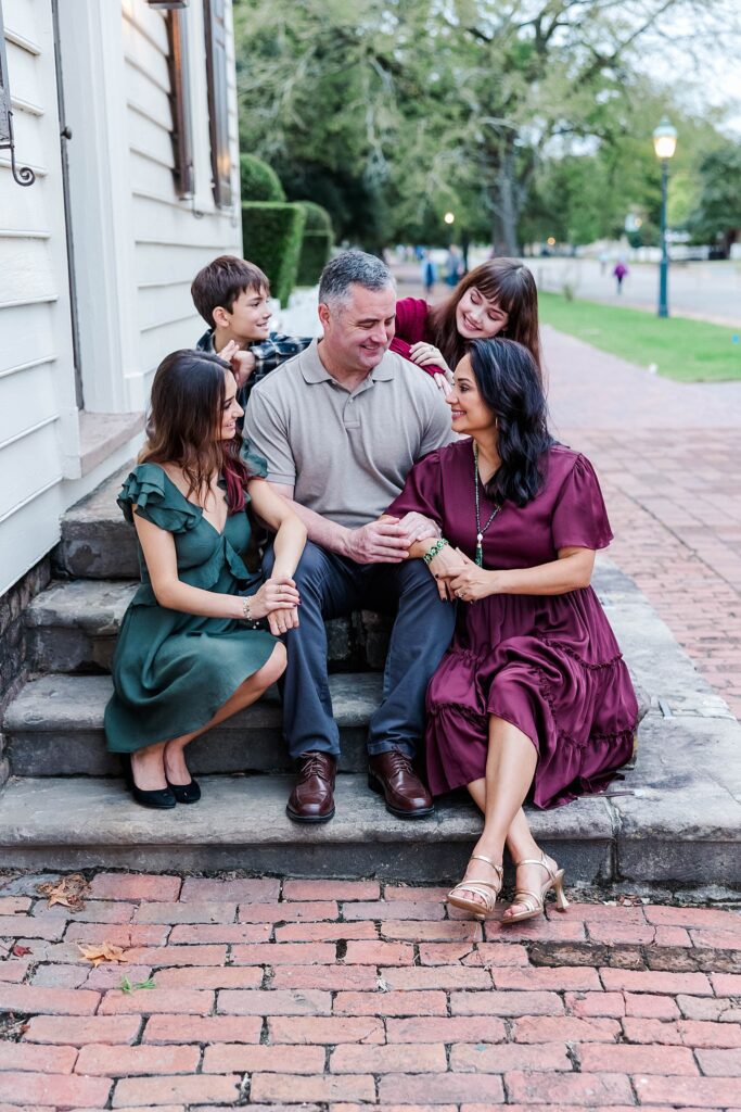 family sits on steps in Colonial Williamsburg laughing for family portrait