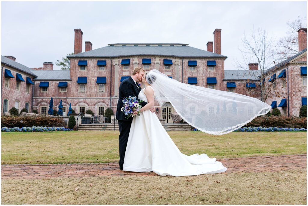 bride and groom kiss with veil blowing in wind behind Williamsburg Inn after their Colonial Williamsburg Wedding