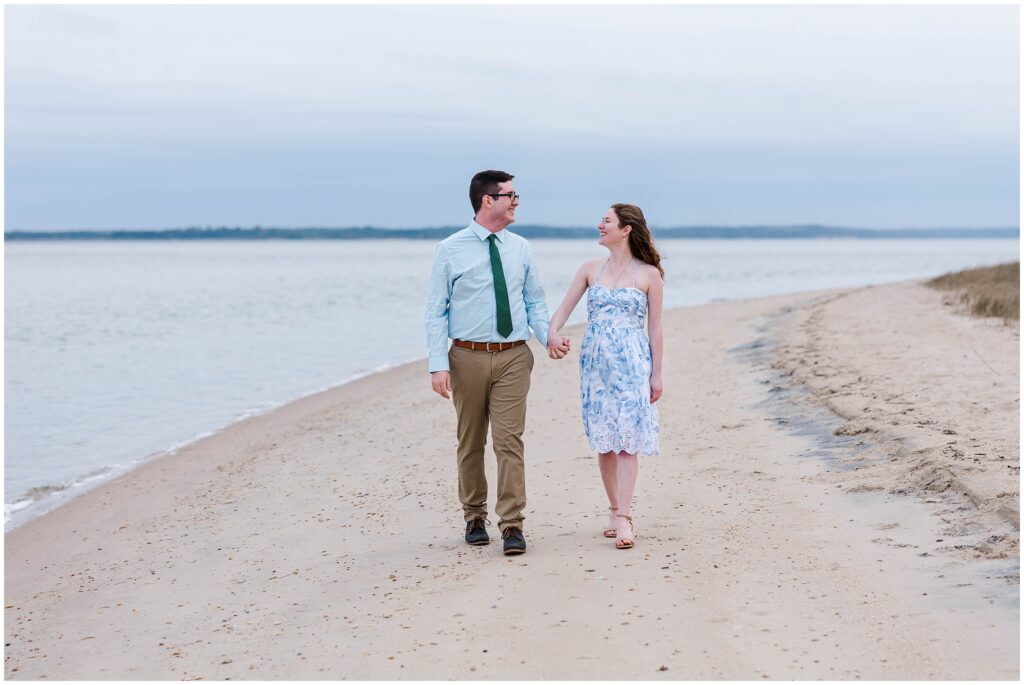 couple walks along beach with glowing serene sky at their beach engagement session in Williamsburg, VA